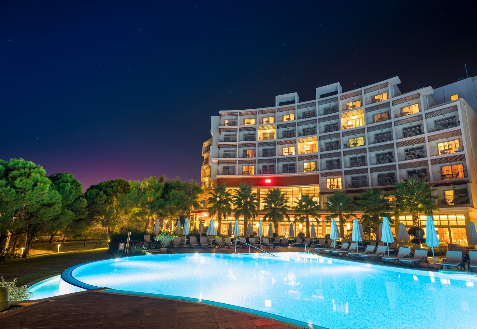 Akra Sorgun Tui Blue Sensatori viesnīcas ēka un izgaismots āra baseins naktī — moderna arhitektūra ar palmām un atpūtas krēsliem