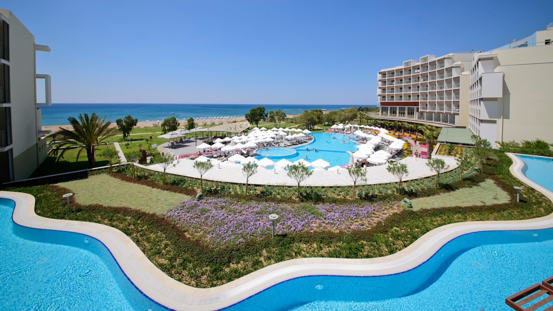 Akra Sorgun Tui Blue Sensatori panorāmas skats uz plašo baseinu kompleksu, dārzu un Vidusjūras piekrasti saulainā dienā
