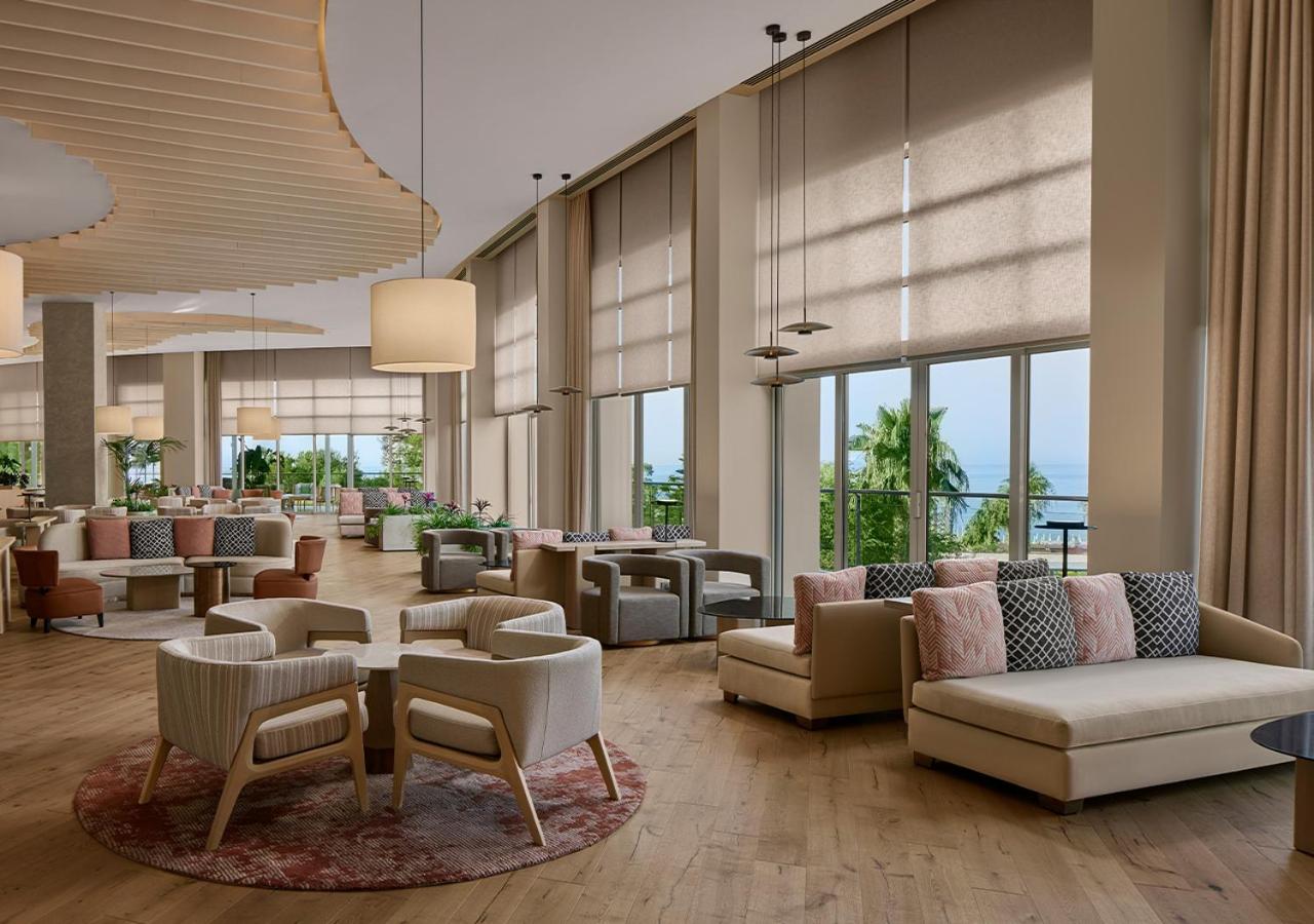 Akra Sorgun Tui Blue Sensatori plašā lobby atpūtas zona ar ērtiem dīvāniem, panorāmas logiem un skatu uz dārzu