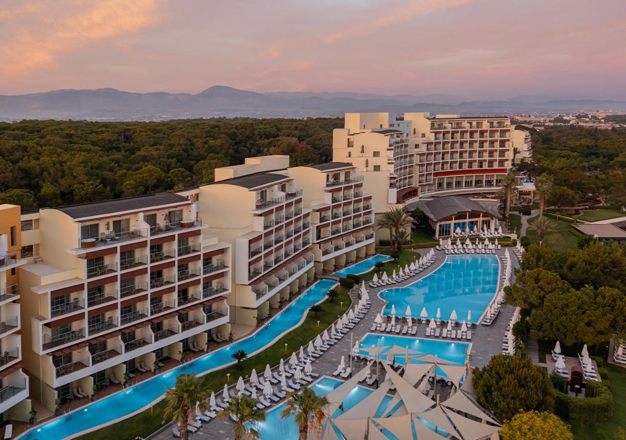 Akra Sorgun Tui Blue Sensatori modernie numuri ar balkoniem un baseinu kompleksu vakara gaismās uz kalnu fona
