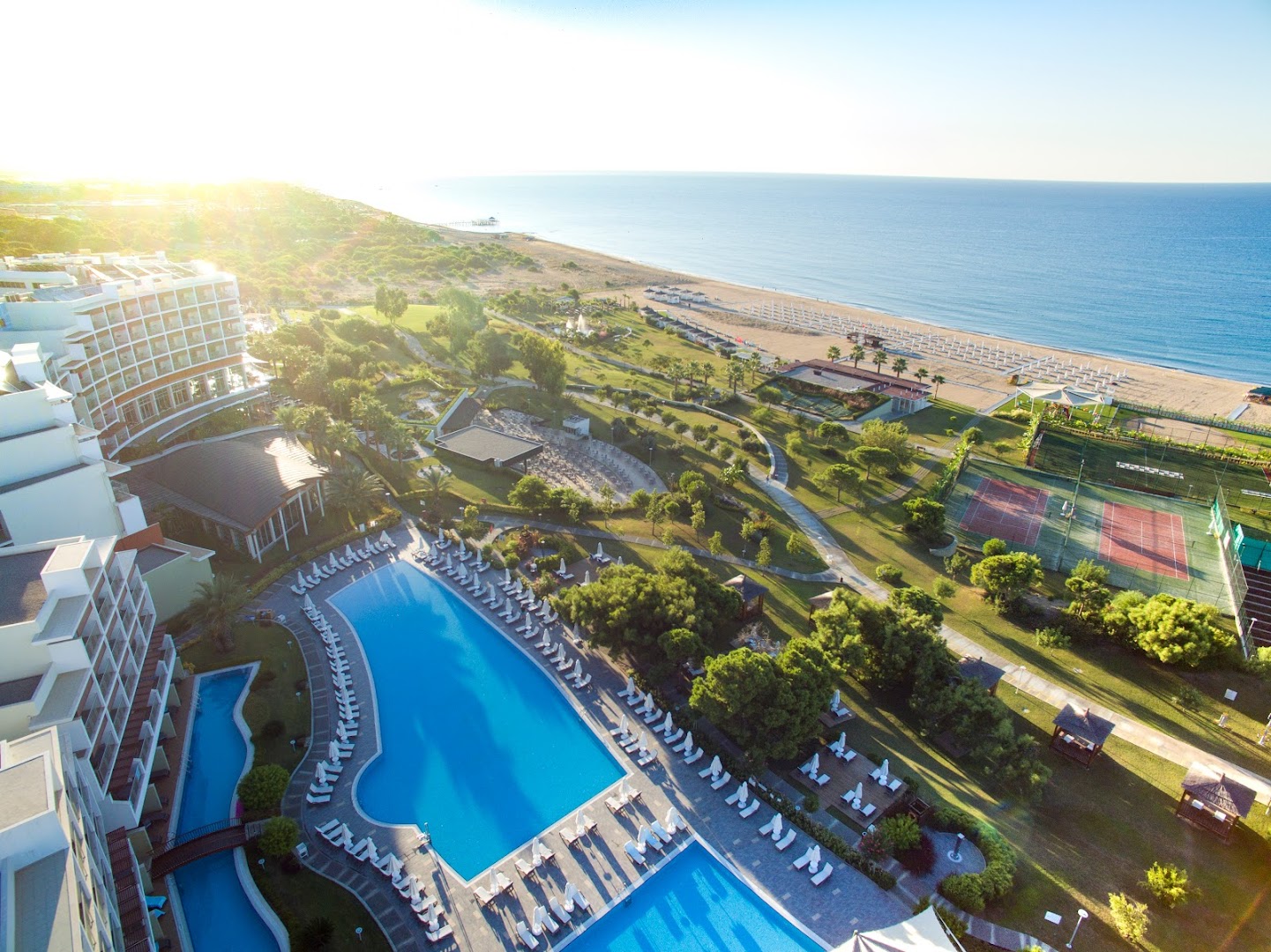 Akra Sorgun Tui Blue Sensatori skats no augšas uz galveno baseinu, dārzu un tenisa kortiem rīta saulē