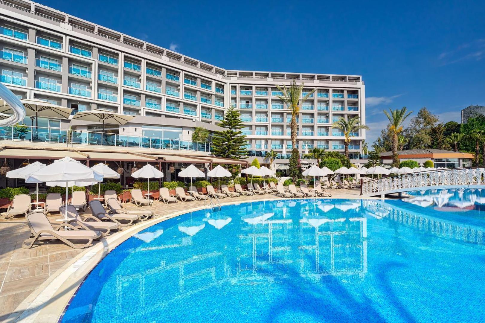 Annabella Diamond Hotel āra baseins dienas laikā ar sauļošanās krēsliem, saulessargiem un viesnīcas korpusa atspulgu ūdenī