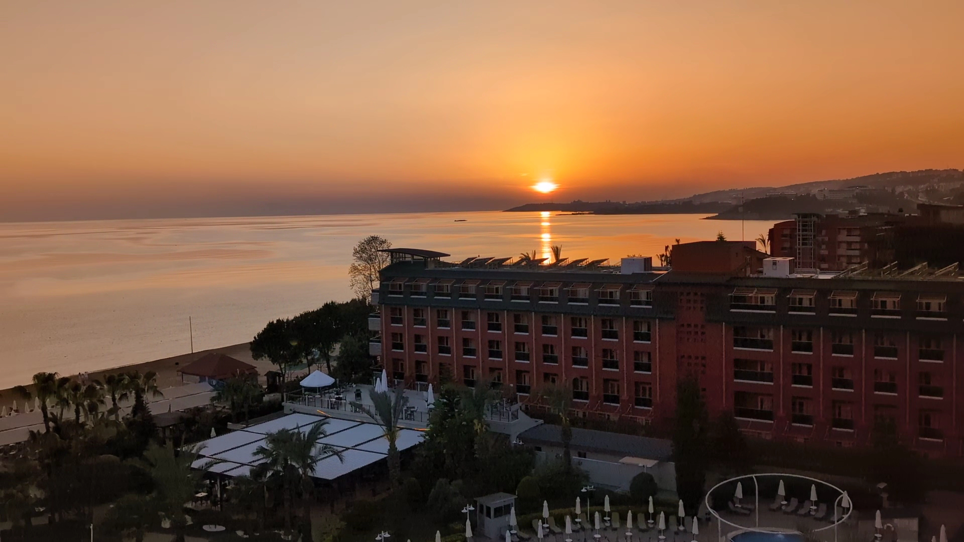 Annabella Diamond Hotel skats no augšas uz jūru saulrietā — viesnīcas ēka piekrastē un oranžas debesis virs horizonta