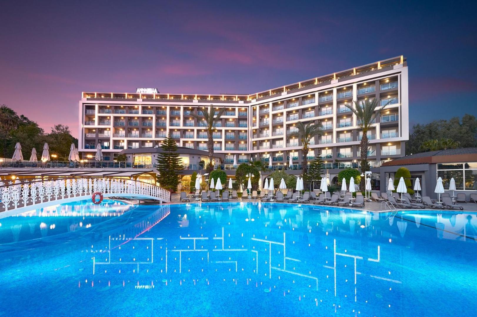 Annabella Diamond Hotel vakara skats uz baseinu ar dekoratīvu tiltiņu un izgaismotiem numuru balkoniem Alanjas piekrastē