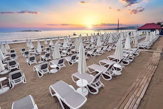 Annabella Diamond Hotel smilšu pludmale Alanjā saulrieta laikā — rindu rindām izkārtoti sauļošanās krēsli un balti saulessargi