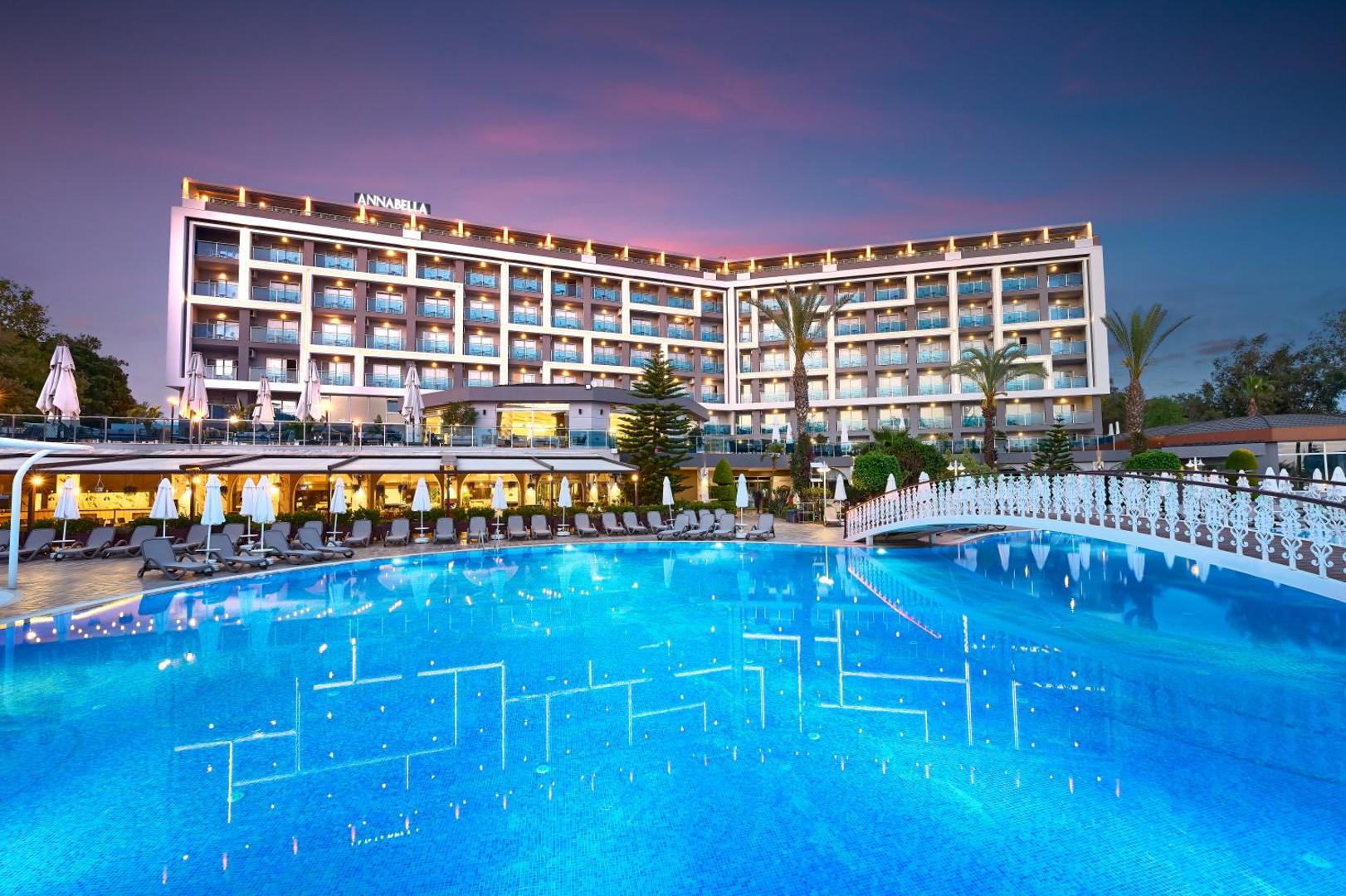 Annabella Diamond Hotel ēkas fasāde un izgaismots āra baseins krēslas stundā Alanjas reģionā, Turcijā — moderna arhitektūra ar balkoniem