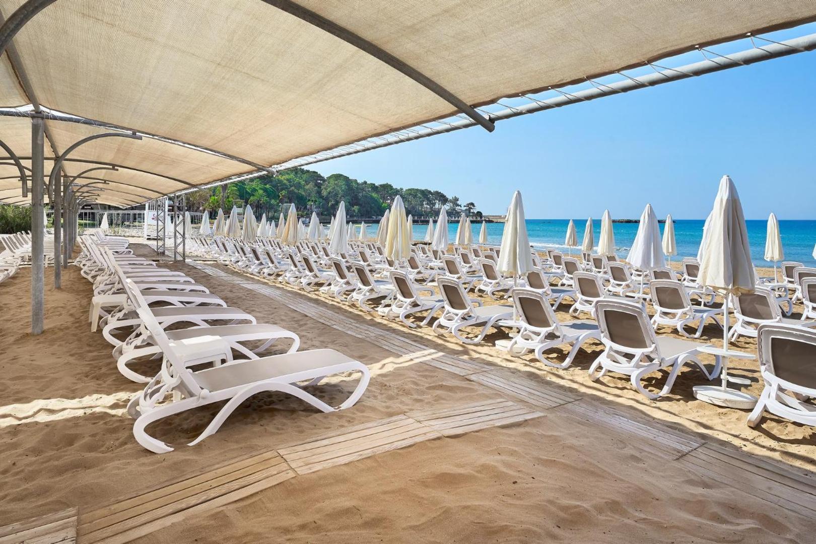 Annabella Diamond Hotel pludmales zona ar nojumi un sauļošanās krēsliem — skats uz zilo jūru un piekrastes mežu