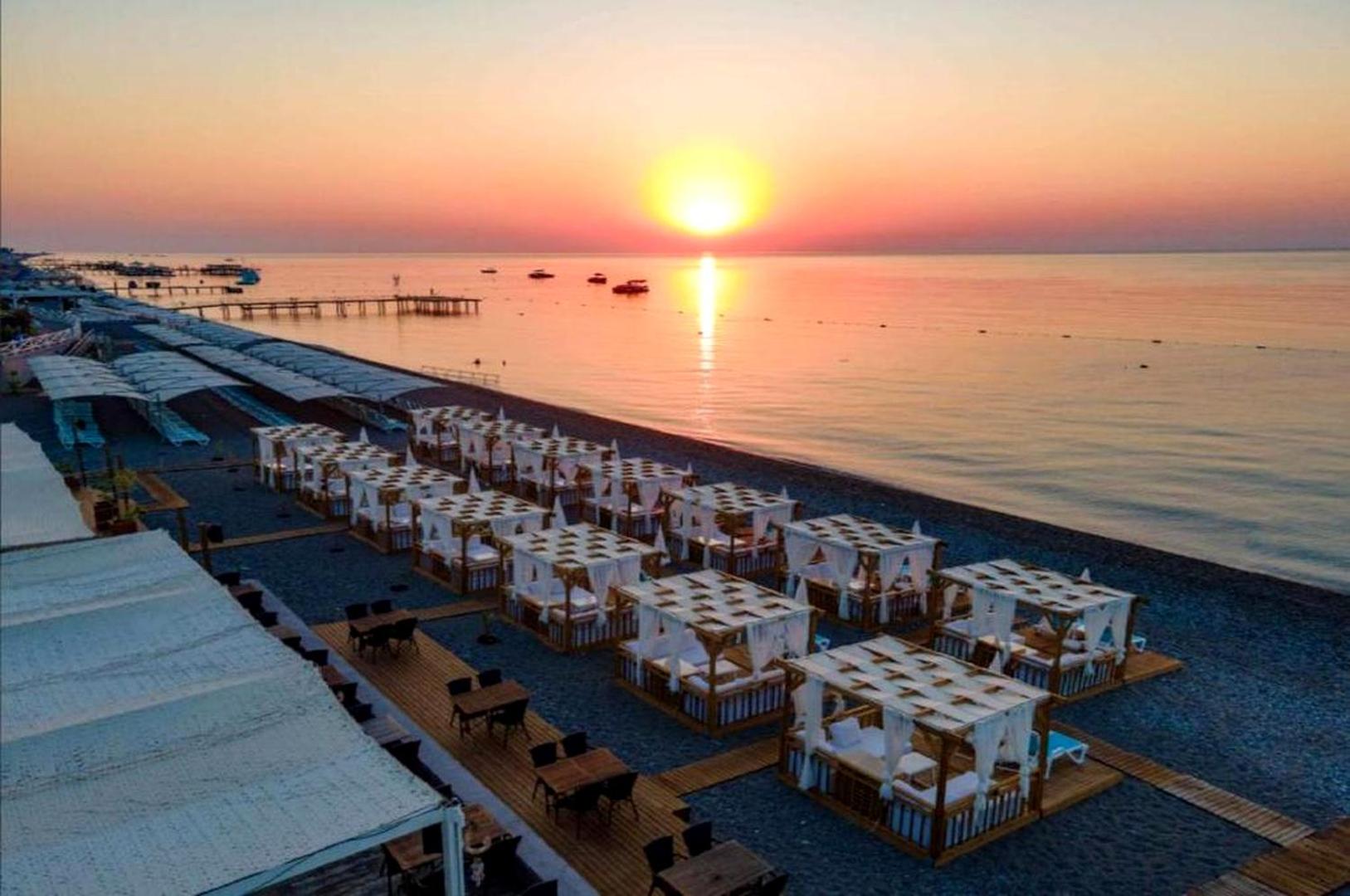 Aerofoto skats uz Club Marco Polo pludmali saulrietā — daudzas baltas nojumes un galdiņi pie jūras krasta