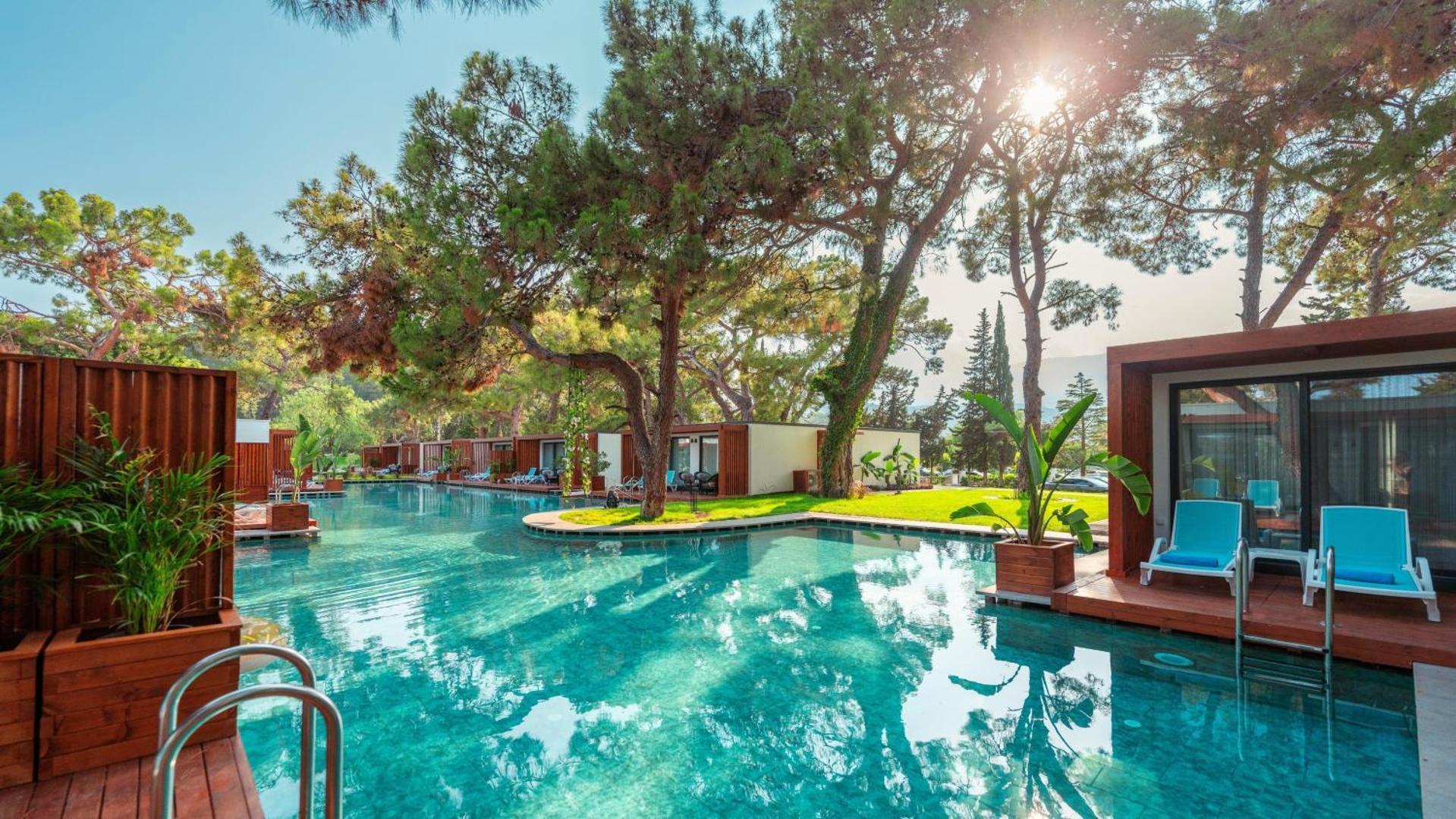 Club Marco Polo moderni bungalo pie baseina Kemeras reģionā — saules apspīdēta atpūtas zona ar koka terasēm un priedēm