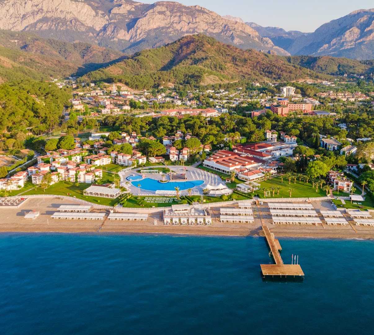 Club Marco Polo kūrorta skats no putna lidojuma saulrietā — redzama pludmales piestātne, baseins un dārza ieskauti numuri