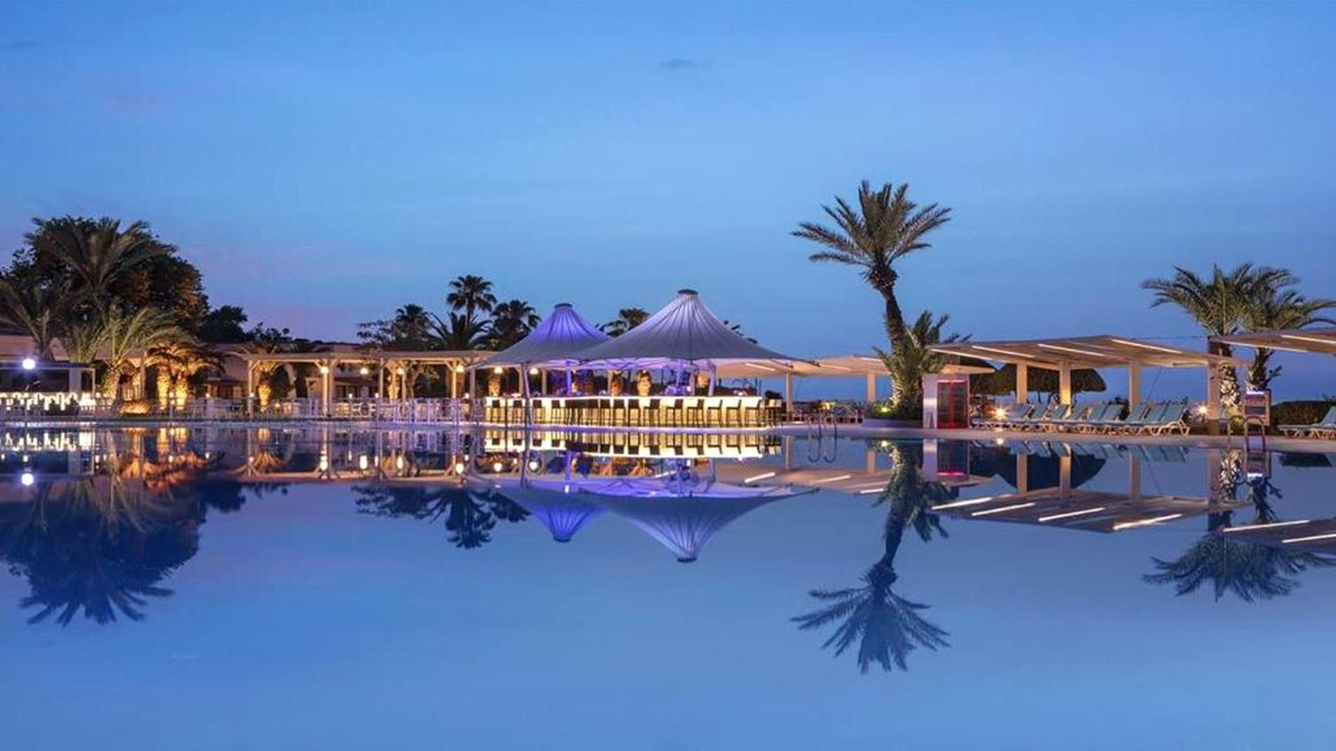 Club Marco Polo vakara skats uz baseinu un izgaismotu bāru teltīs — palma un atspulgi ūdenī Kemeras piekrastē