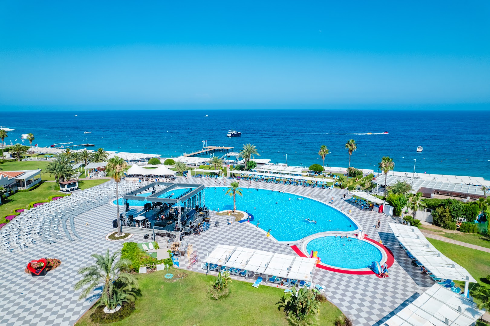 Club Marco Polo panorāmas skats uz lielo baseinu, skatuvi un Vidusjūras piekrasti — plaša atpūtas zona ar sauļošanās krēsliem
