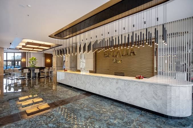 Eftalia Blue Hotel plašā un modernā recepcijas zāle ar marmora apdari, dizaina gaismekļiem un viesnīcas logo pie sienas