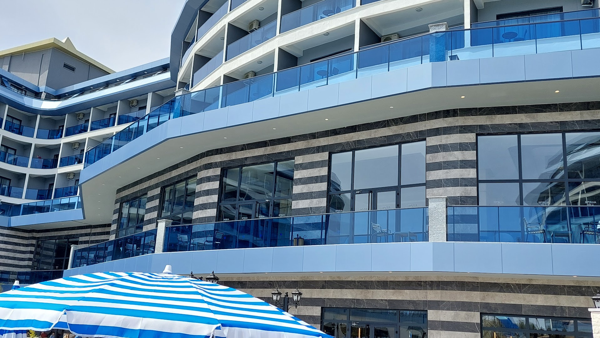 Eftalia Blue Hotel modernā fasāde ar stiklotiem balkoniem un svītrainu saulessargu priekšplānā saulainā dienā Turcijas piekrastē