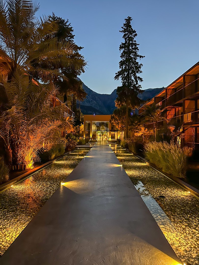 Viesnīcas Balmy Beach Resort Kemer izgaismotais centrālais celiņš vakarā ar dekoratīviem baseiniem un kalnu siluetu tālumā