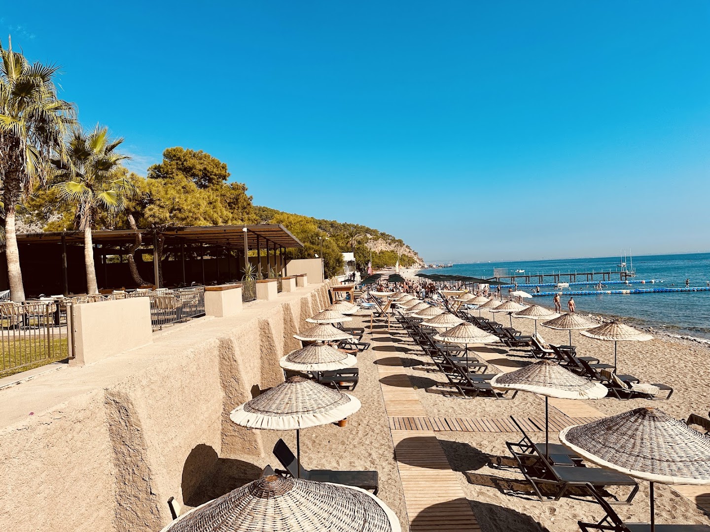 Balmy Beach Resort Kemer privātā pludmale ar saulessargiem, sauļošanās krēsliem un koka laipu — skats uz jūru un kalniem