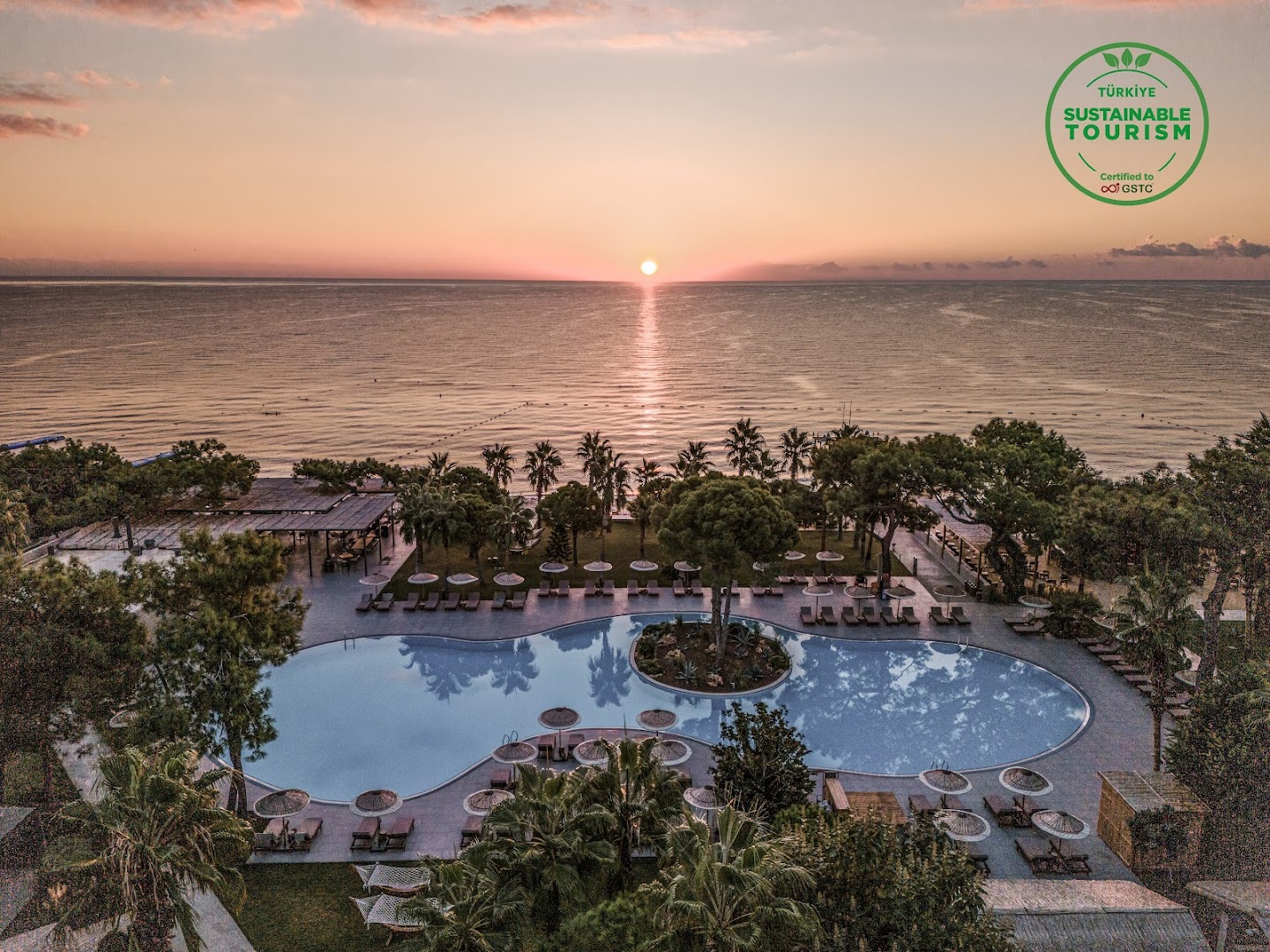 Saulriets pār Vidusjūru pie Balmy Beach Resort Kemer — skats uz pludmali, baseinu un palmām vakara saules gaismā