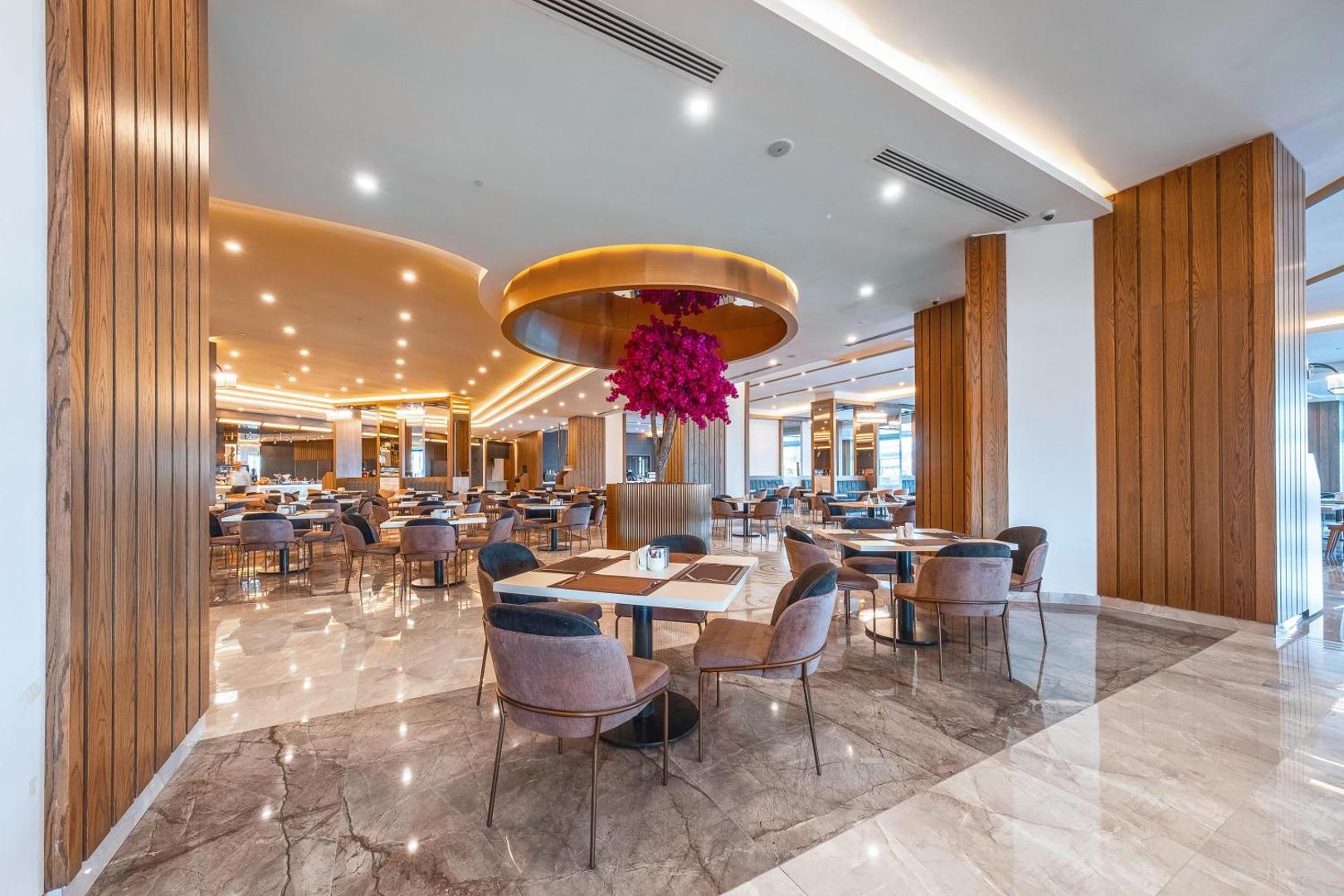 Lago Hotel restorāna zāle ar elegantiem galdiņiem, mīkstiem krēsliem un dekoratīvu ziedošu koku telpas centrā