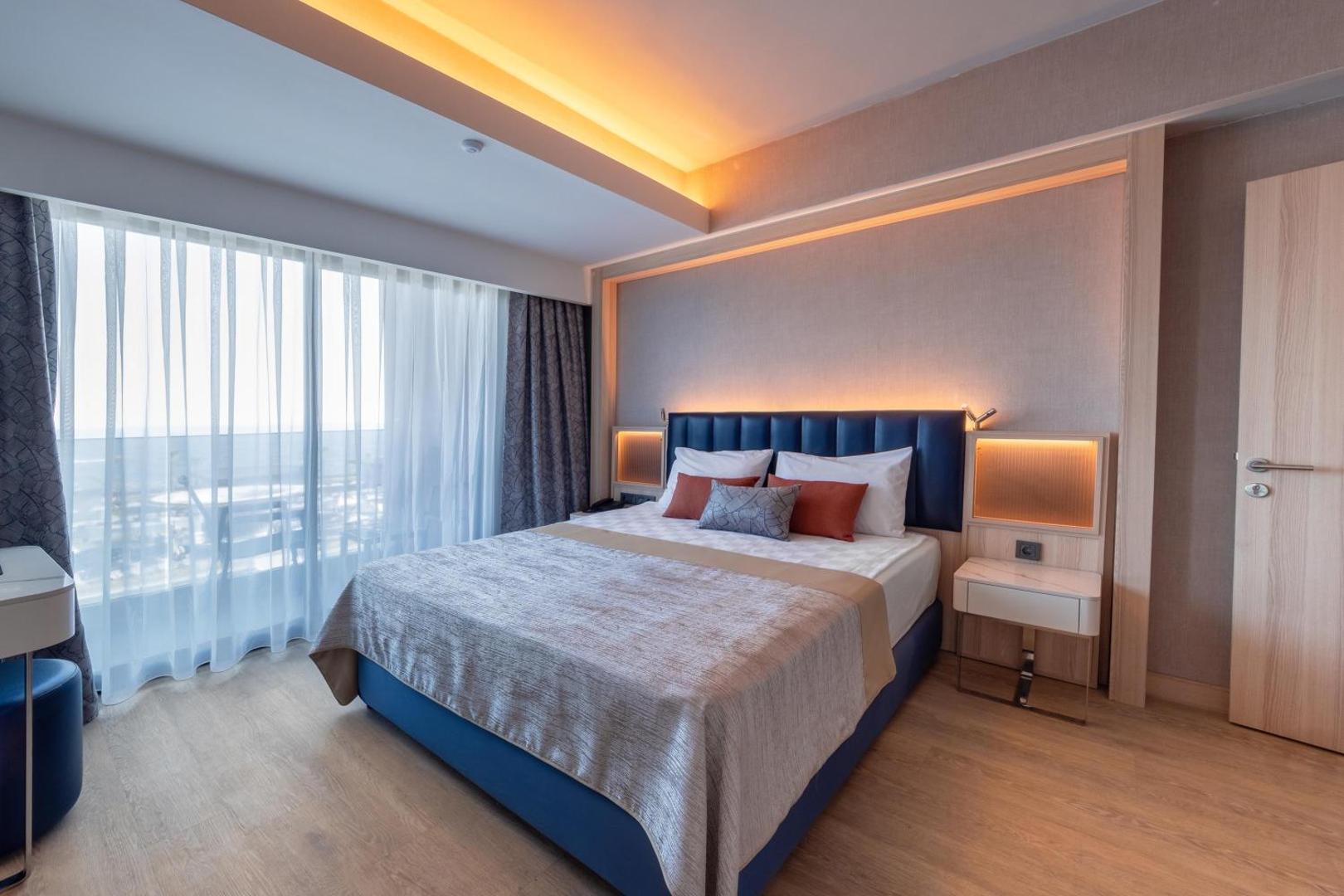 Lago Hotel moderns divvietīgs numurs ar gaišu koka grīdu, zilu gultas galvgalī un lielu logu uz balkonu Turcijā