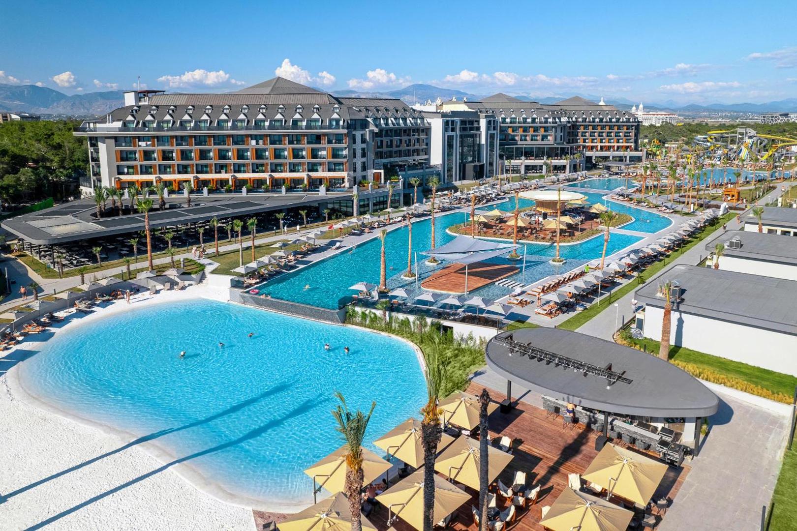 Panorāmas skats uz Lago Hotel kompleksu Sidē — modernas ēkas, plaši baseini un akvaparks fonā zem skaidrām debesīm