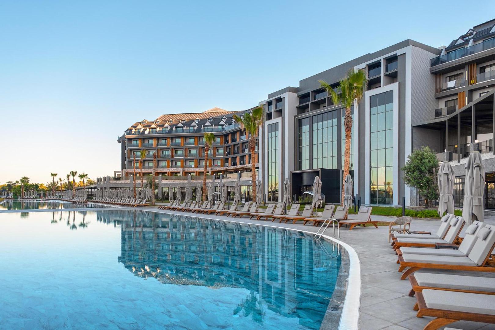 Lago Hotel modernā fasāde un lielais āra baseins saulrieta gaismā Sidē, Turcijā — atpūtas zona ar ērtiem zviļņiem un palmām