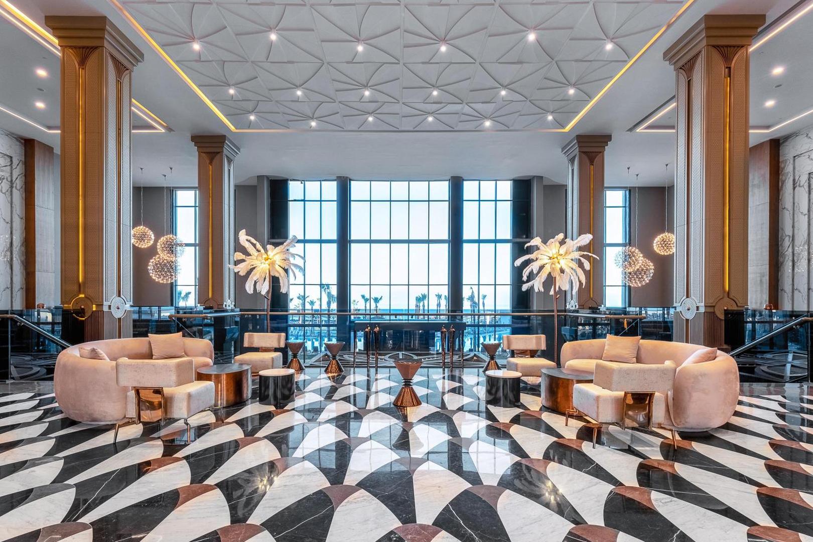 Lago Hotel greznais vestibils ar marmora grīdu, dizaina mēbelēm un lieliem logiem Sidē — eleganta atpūtas zona viesnīcas interjerā