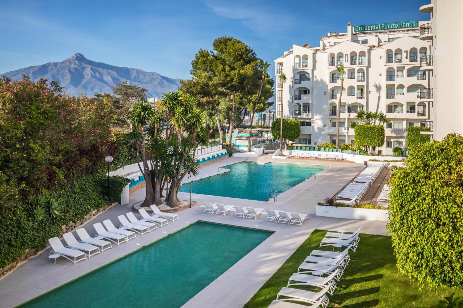 Occidental Puerto Banus viesnīcas teritorija ar diviem āra baseiniem, sauļošanās krēsliem un skatu uz kalniem Marbeljas piekrastē