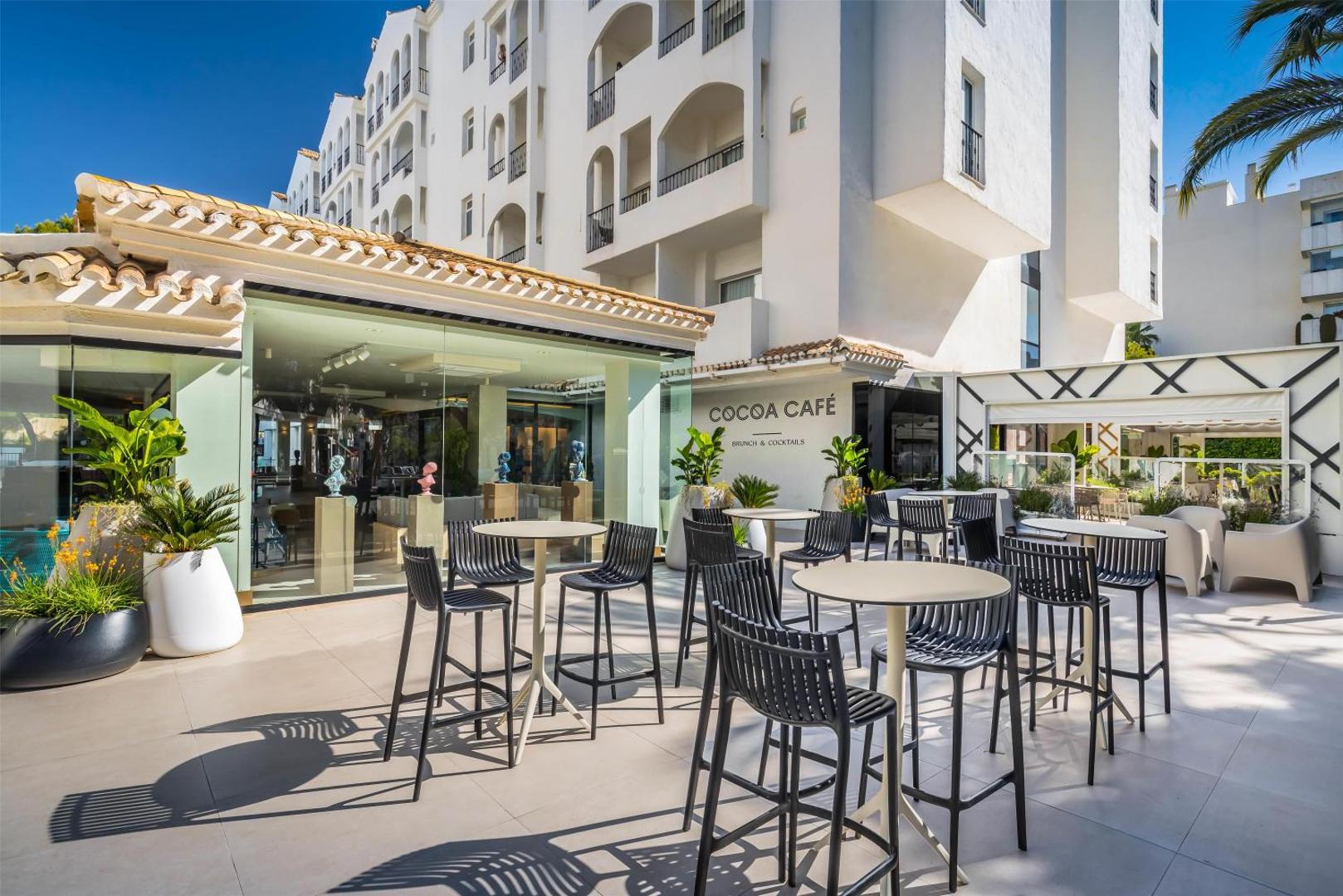 Occidental Puerto Banus viesnīcas Cocoa Café āra terase ar moderniem bāra krēsliem un galdiņiem saulainā pagalmā pie baltas ēkas fasādes