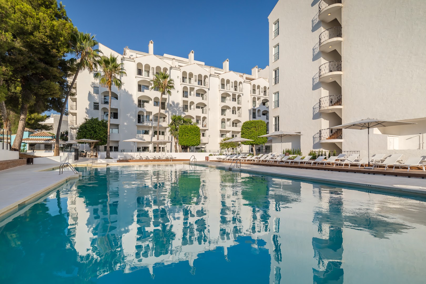 Occidental Puerto Banus lielais āra baseins ar debess atspulgu ūdenī, baltu ēkas fasādi un palmām Marbeljā