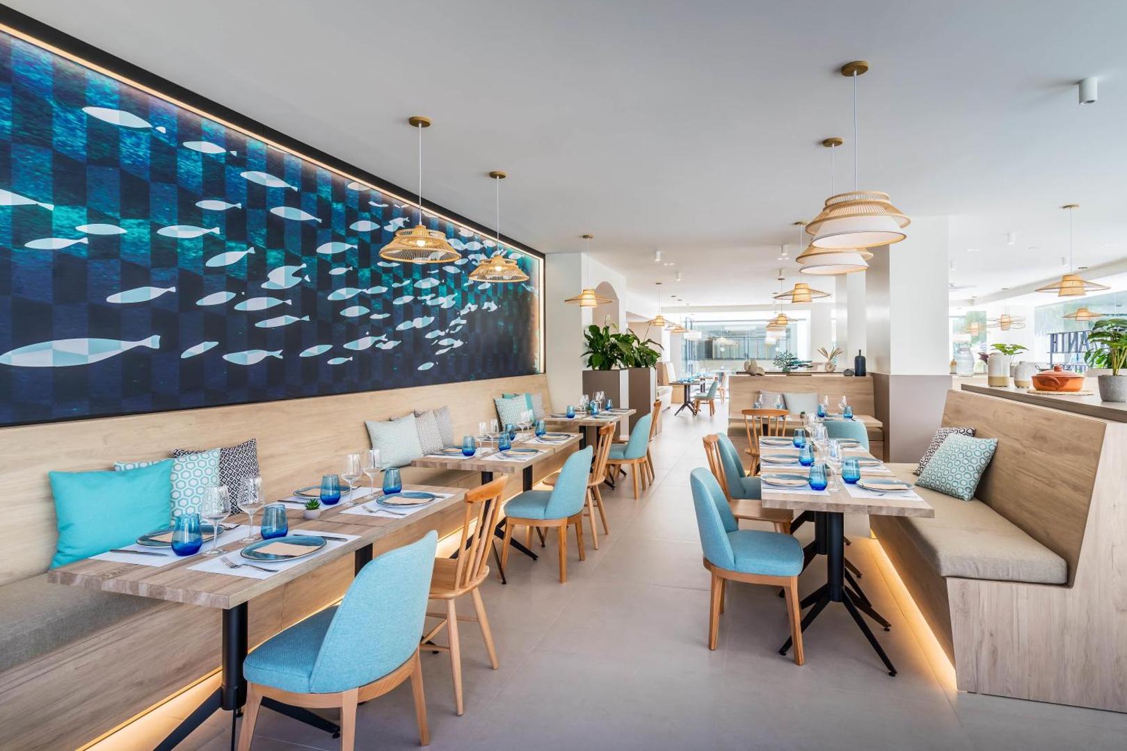 Gaišs Occidental Puerto Banus restorāns ar ziliem krēsliem, koka galdiem un sienas dekoru ar zivju motīviem Marbeljas kūrortā