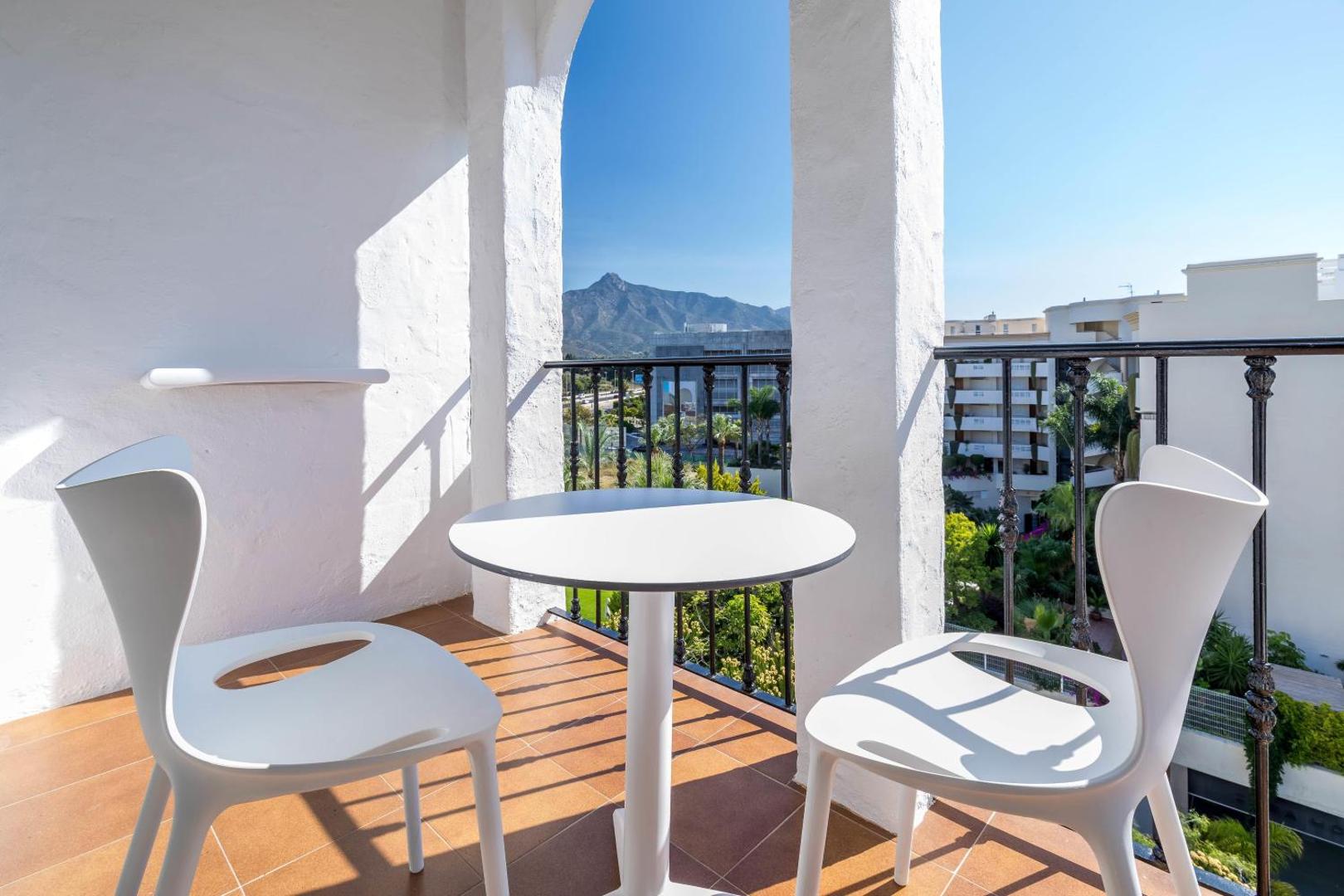 Occidental Puerto Banus numura balkons ar baltām mēbelēm un skatu uz kalniem un zaļojošu dārzu saulainā dienā