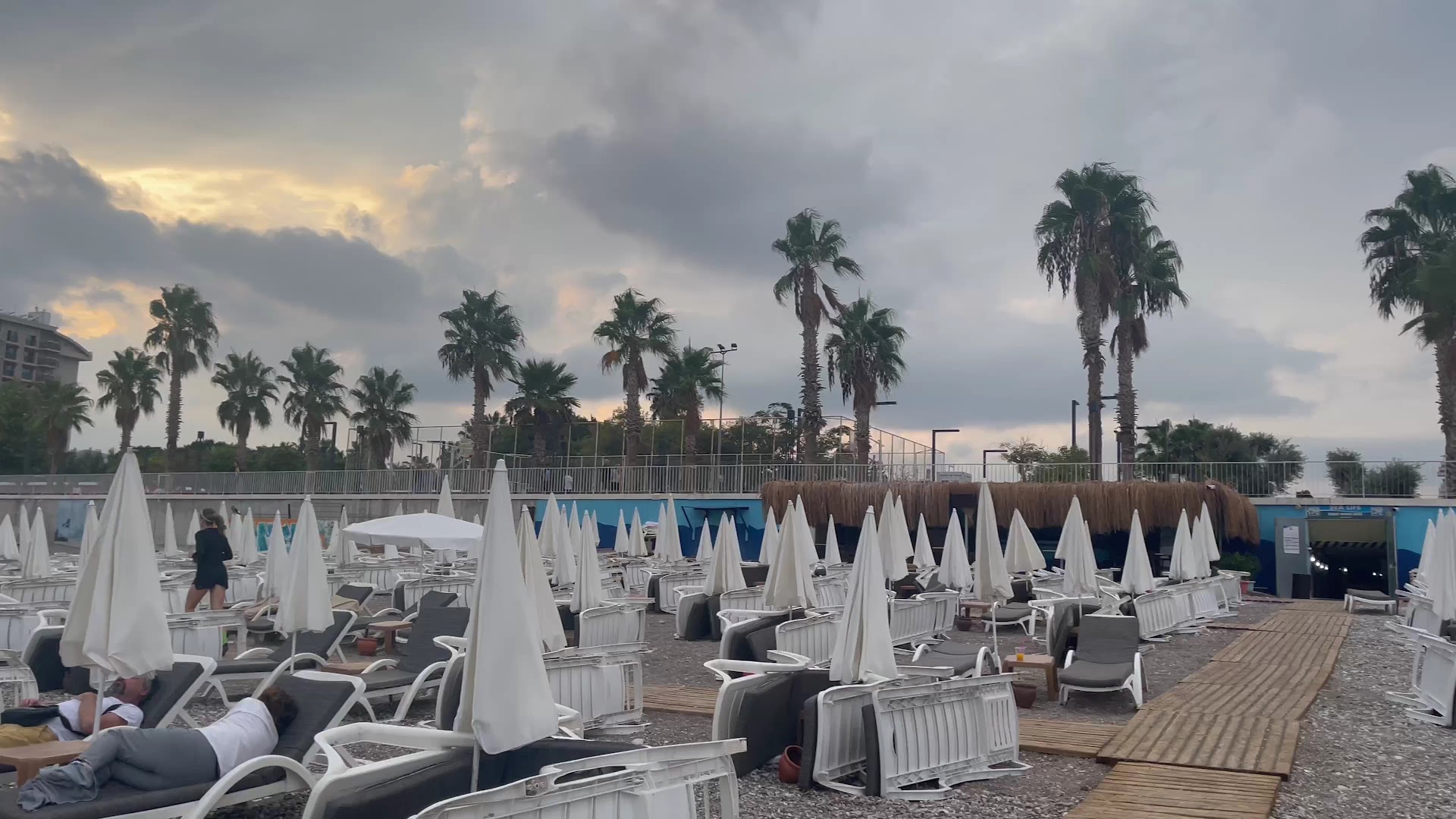 Sealife Family Resort Hotel pludmales zona ar daudziem saulessargiem, atpūtas krēsliem un palmām mākoņainā pievakarē