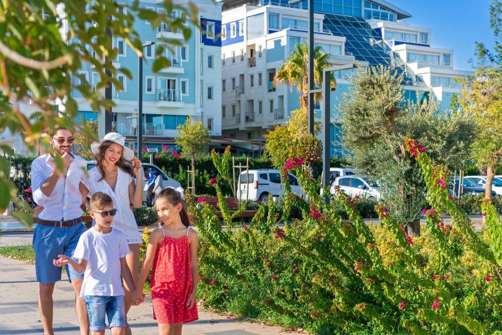 Ģimene ar diviem bērniem pastaigājas pie Sealife Family Resort Hotel — fonā redzama viesnīcas ēka, ziedoši krūmi un palmas