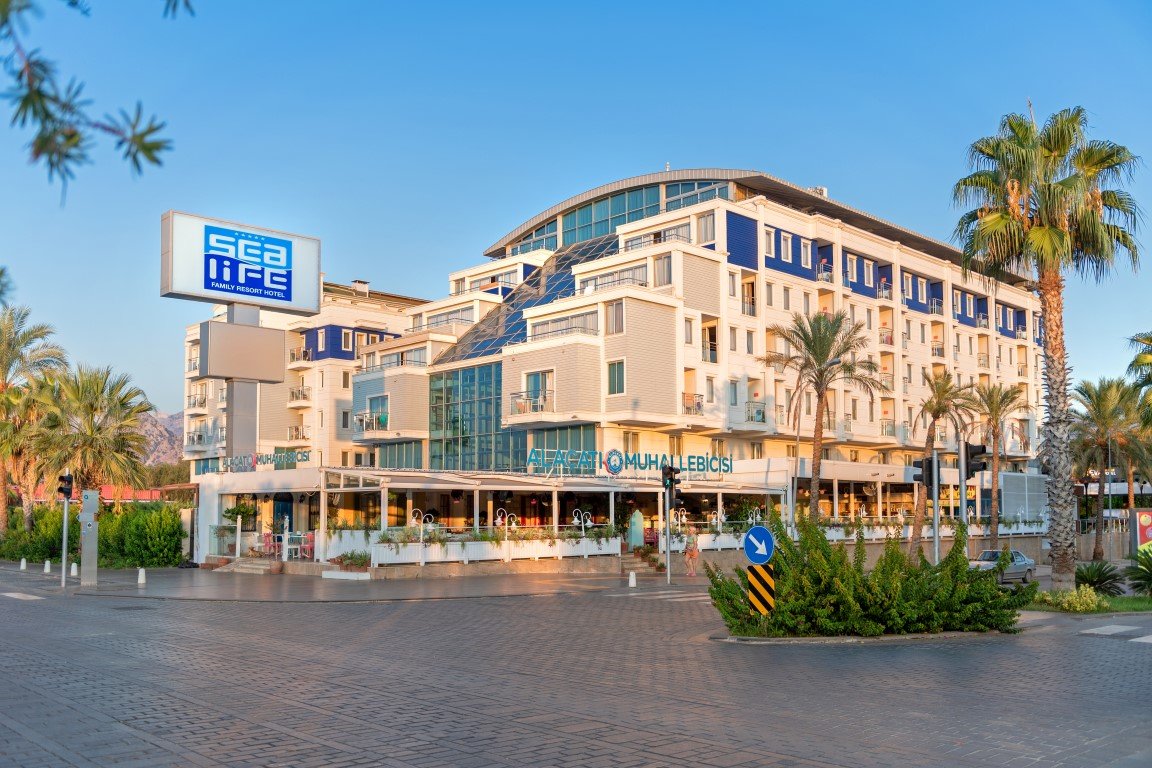 Sealife Family Resort Hotel fasāde un ieeja no ielas puses saulrietā — redzams viesnīcas logo, restorāna terase un apkārtējās palmas