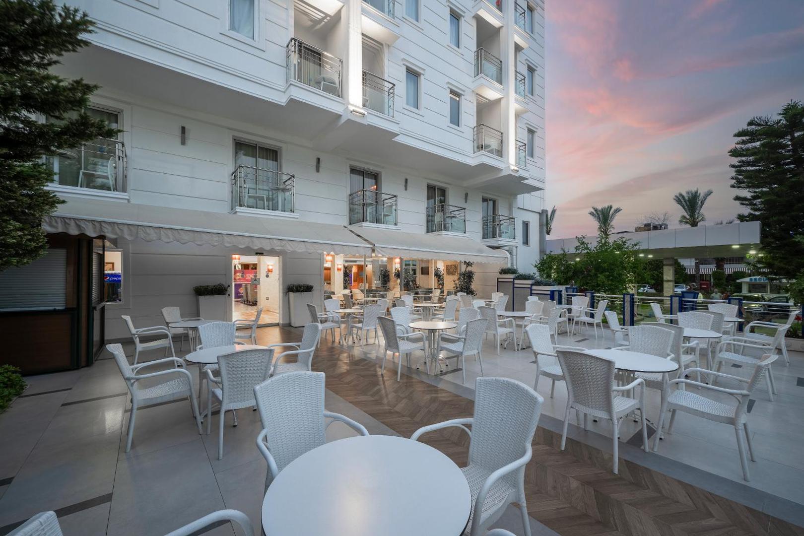 Sealife Family Resort Hotel āra terase ar baltiem krēsliem un galdiem pie viesnīcas fasādes saulrieta laikā Antālijā