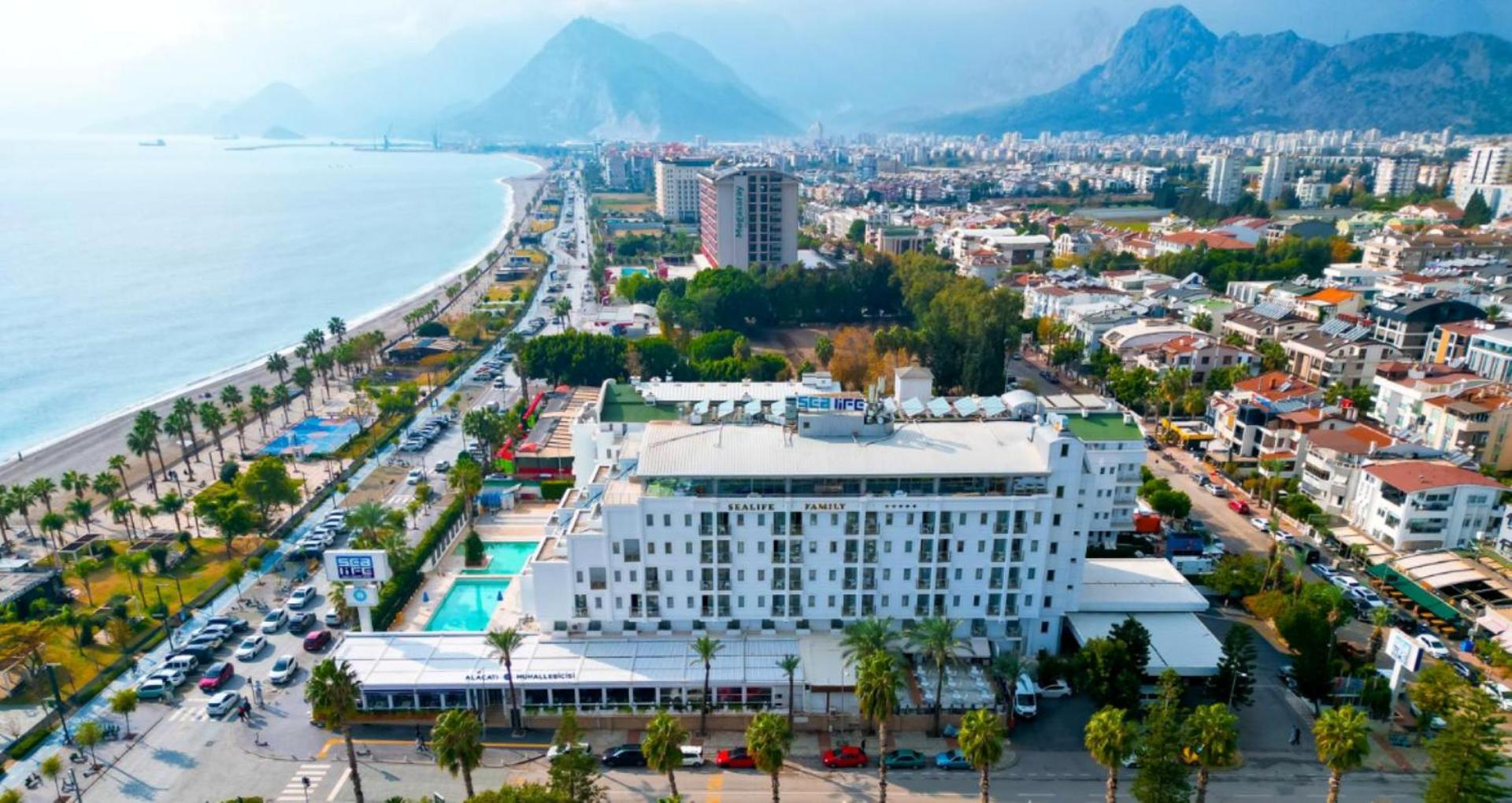 Sealife Family Resort Hotel panorāmas skats no putna lidojuma Antaljā — viesnīcas ēka, āra baseini un Konjaliti pludmales piekraste ar kalniem fonā