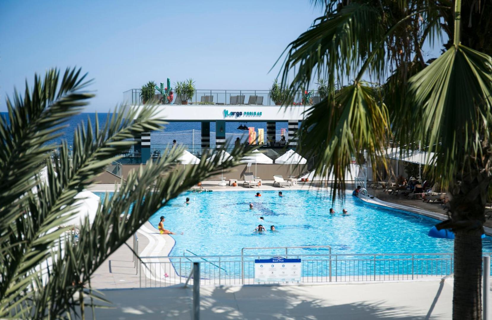 Skats caur palmu lapām uz Sailor's Beach Club āra baseinu un baseina bāru pie Vidusjūras krasta Kemerā