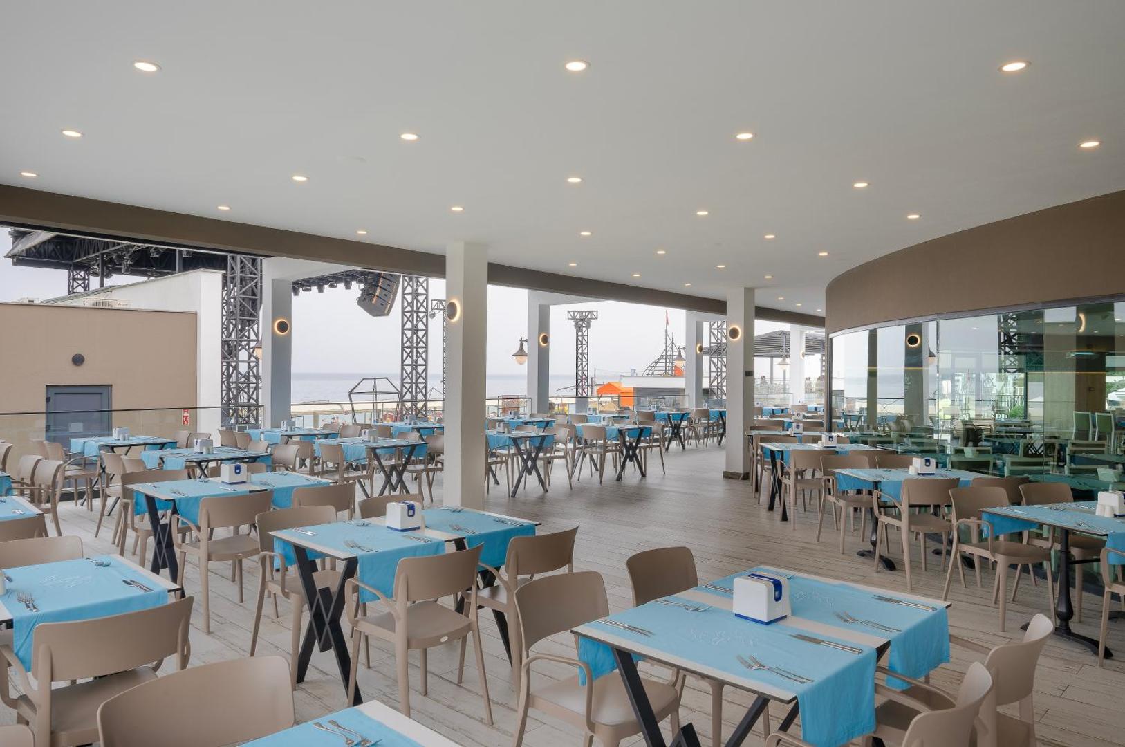Sailor's Beach Club āra restorāns ar gaiši ziliem galdiem un skatu uz jūru un pasākumu skatuvi
