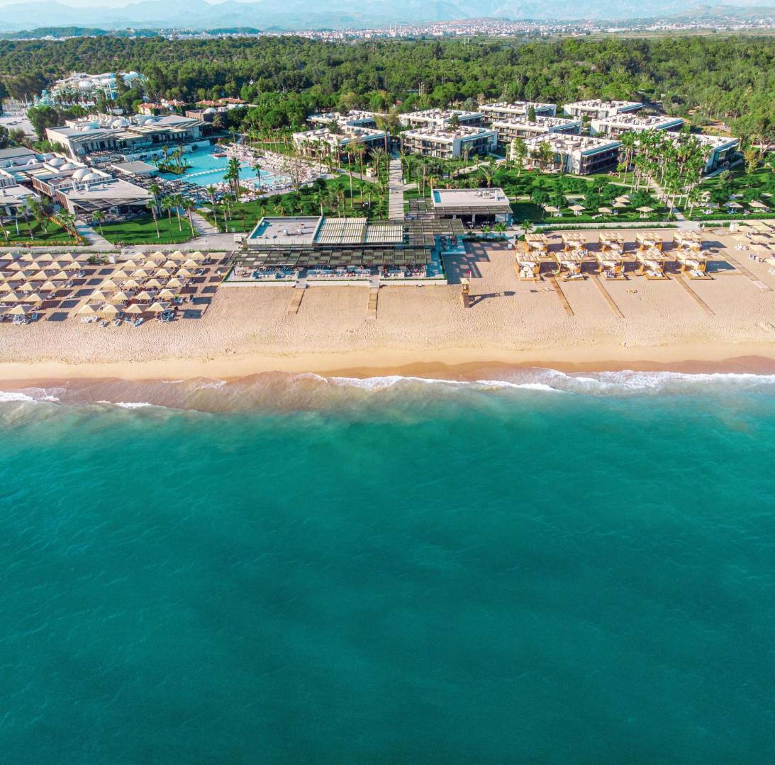 Paloma Orenda viesnīcas panorāmas skats no augšas Sidē, Turcijā — plaša smilšu pludmale, tirkīza jūra un kūrorta teritorija ar baseiniem