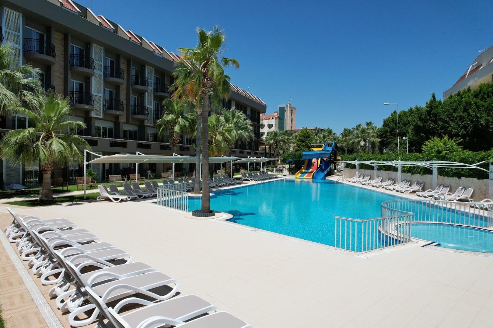 Camyuva Beach Hotel plašā sauļošanās zona pie baseina ar daudziem zviļņiem, saulessargiem un palmām Kemeras kūrortā