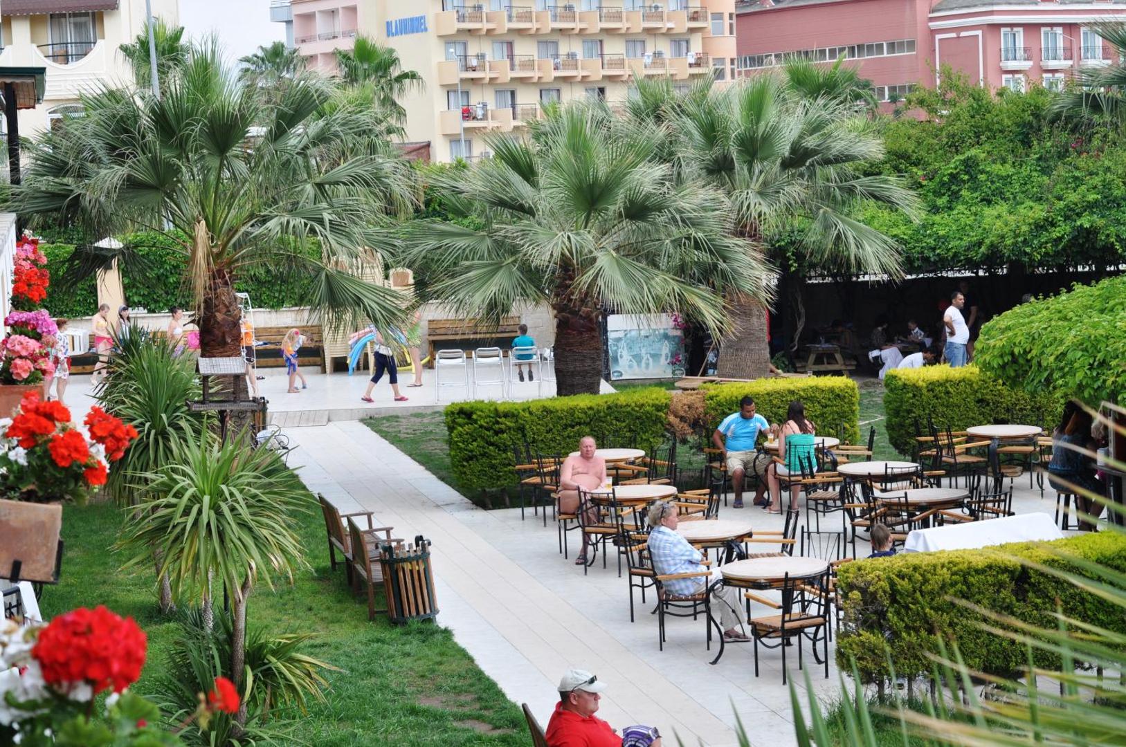 Camyuva Beach Hotel dārza terase ar galdiņiem un palmām — atpūtas zona starp viesnīcas ēkām Kemeras piekrastē