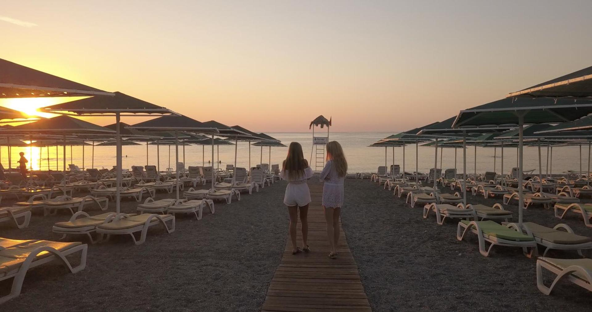 Saulriets Camyuva Beach Hotel privātajā pludmalē — divas sievietes vēro jūru starp saulessargiem un sauļošanās krēsliem