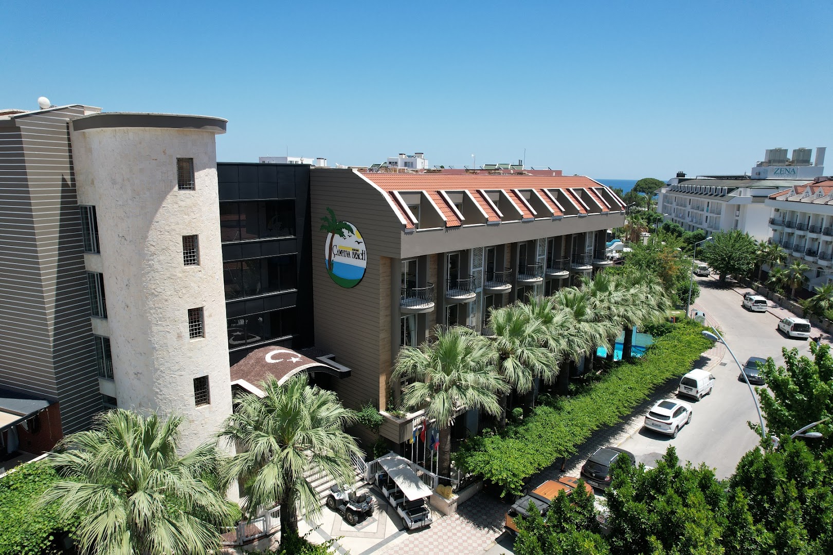 Camyuva Beach Hotel galvenā ieeja un fasāde ar palmām, modernu arhitektūru un viesnīcas logo Kemeras piekrastē.