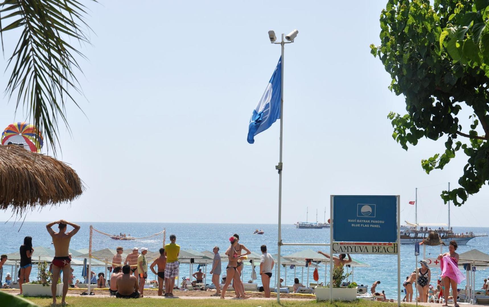 Camyuva Beach Hotel pludmale ar Zilo karogu — skats uz jūru, volejbola tīklu un atpūtniekiem saulainā dienā