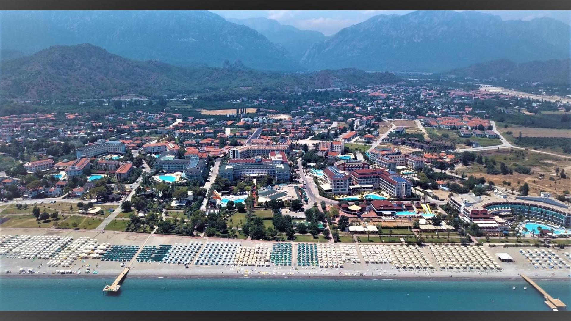 Panorāmas skats uz Čamjuvas kūrortu, plašo oļu pludmali ar saulessargiem, piestātnēm un kalnu grēdu tālumā.