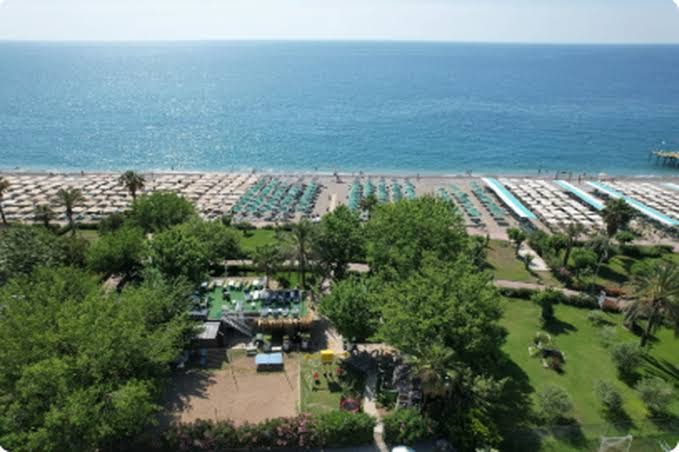 Aerofoto skats uz Camyuva Beach Hotel plašo pludmali ar saulessargiem un zaļo dārza teritoriju piekrastē