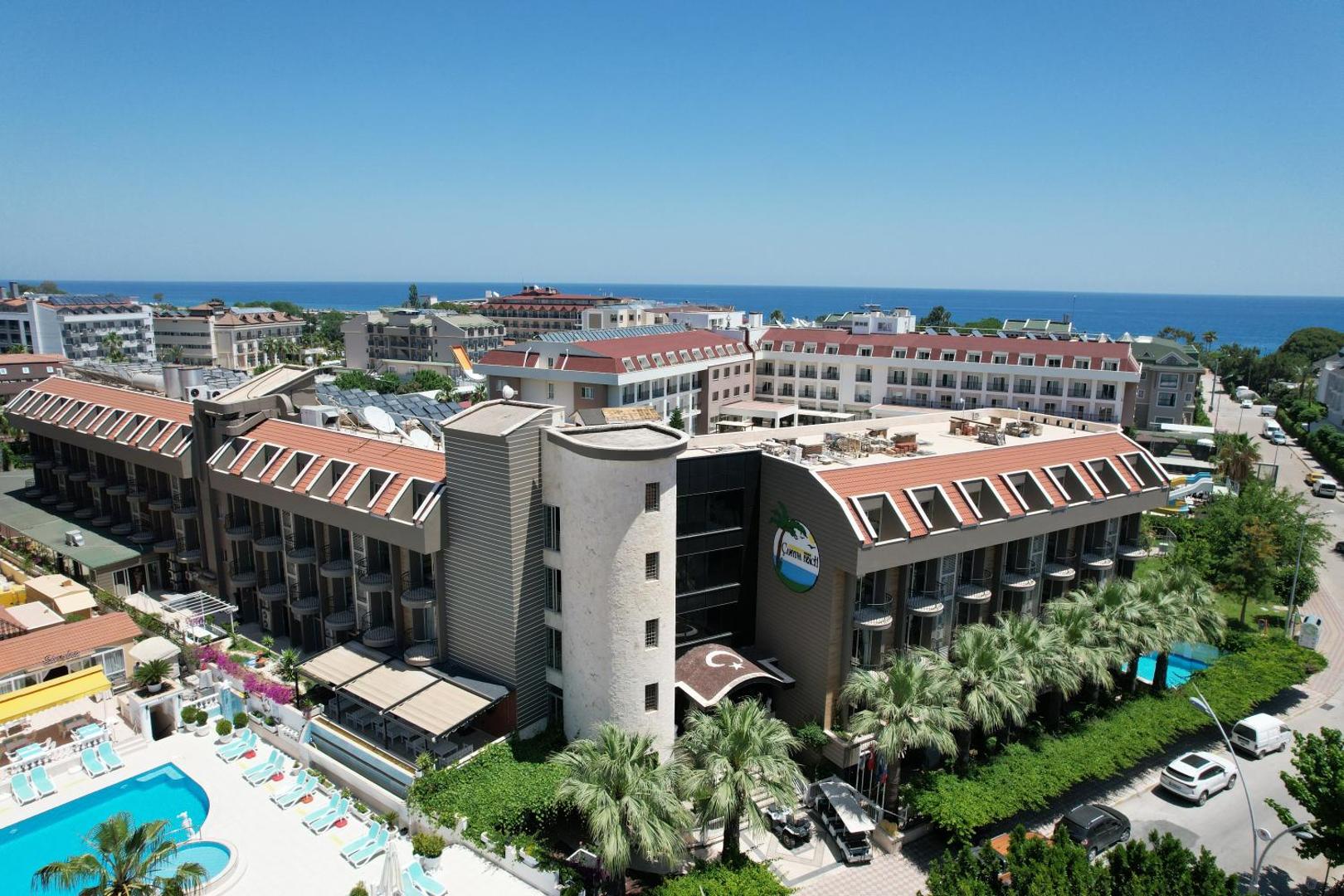 Camyuva Beach Hotel ēku komplekss no putna lidojuma ar skatu uz Vidusjūru, baseinu zonu un apkārtējo pilsētas ainavu.