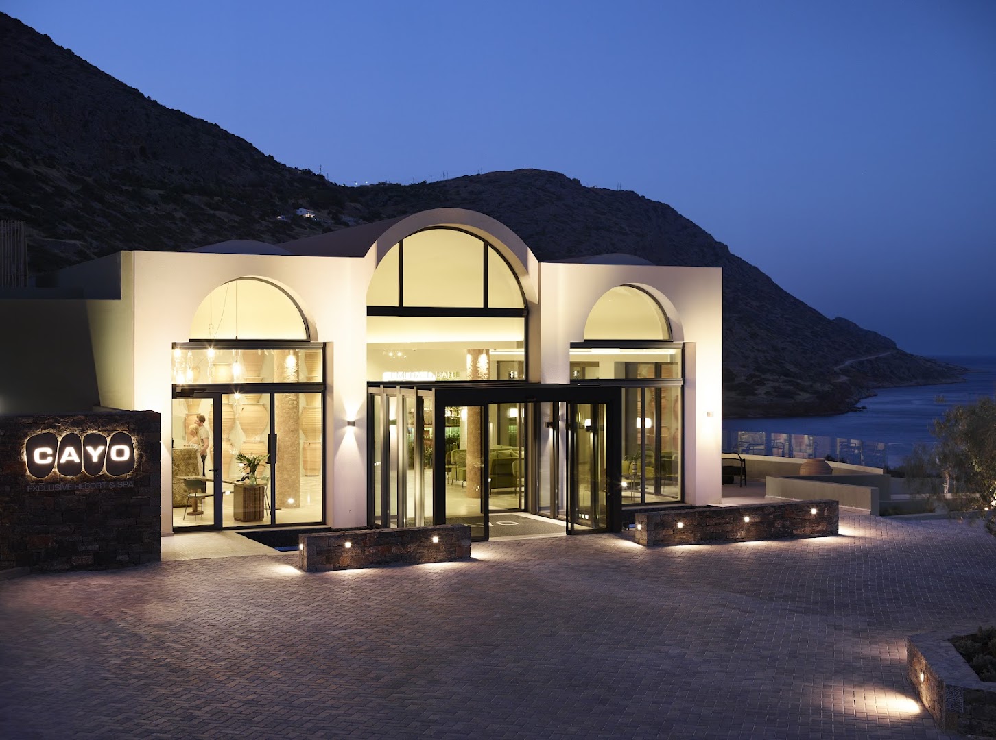 Cayo Exclusive Resort and Spa galvenā ieeja vakarā Elundā, Krētā — apgaismota moderna arhitektūra ar arkveida logiem un akmens sienām