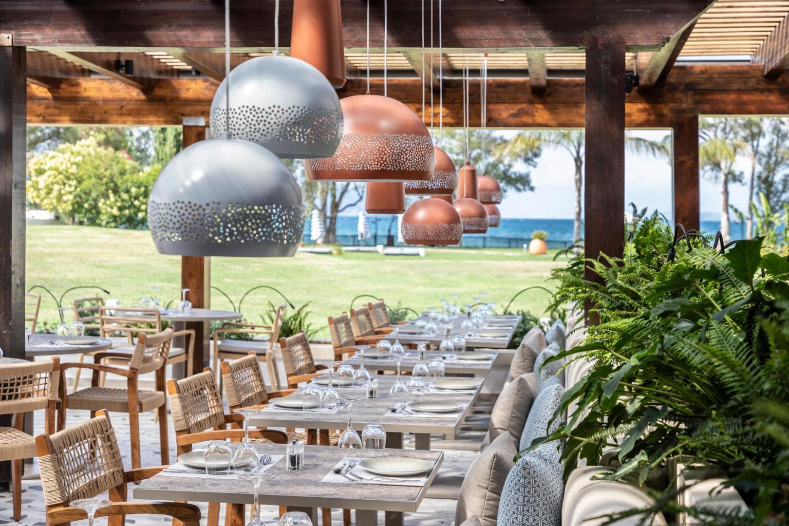 Kerkyra Blue Hotel & Spa āra restorāna terase ar skatu uz jūru Korfu salā — eleganti klāti galdi un dizaina gaismekļi