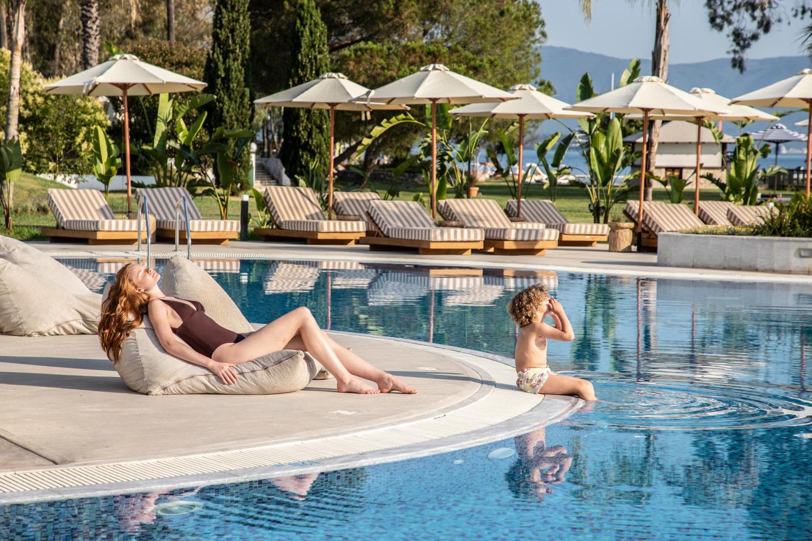 Atpūta pie Kerkyra Blue Hotel & Spa baseina, kur sieviete sauļojas sēžammaisā un bērns spēlējas pie ūdens malas palmu ieskautā teritorijā.